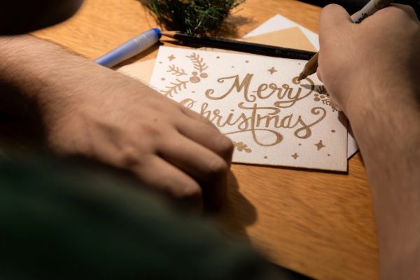 Person writing a festive "Merry Christmas" card with decorative designs.