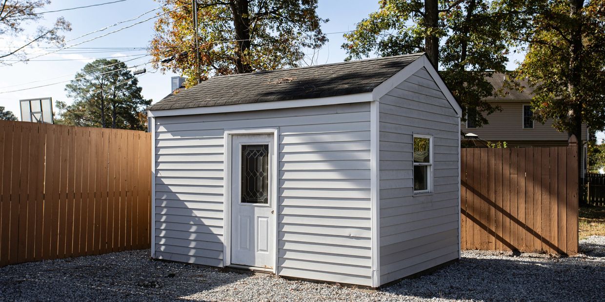 Small white garden shed with gravel ground and wooden fence.