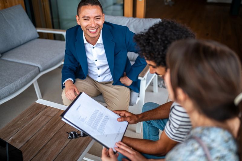 A cheerful diverse couple sits with a professional agent, attentively reviewing a home purchase contract, as they prepare to make an informed decision on their new property.