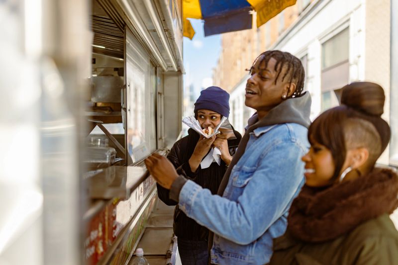 Three friends enjoy ordering food from a food truck in urban setting during the day.