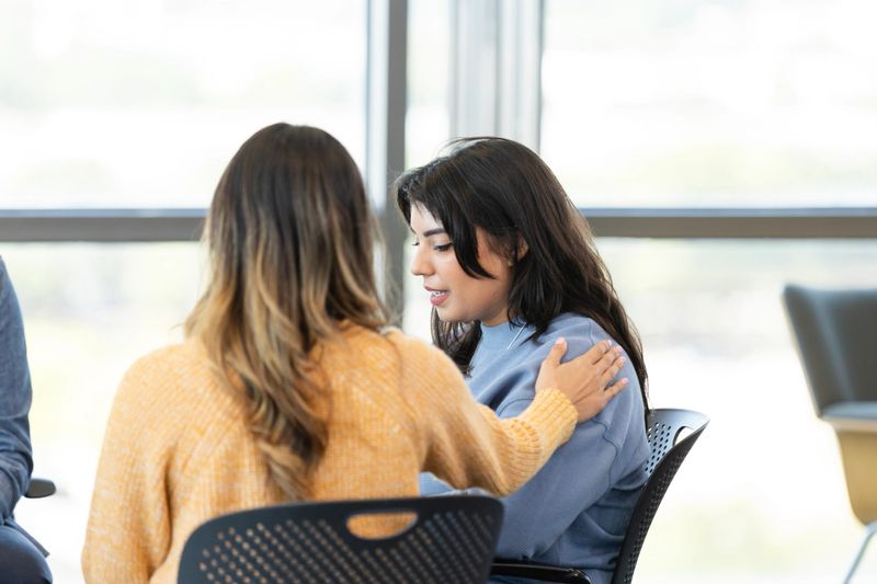 An emotional young woman shares something difficult during a group therapy session. Her friend consoles her as she shares.