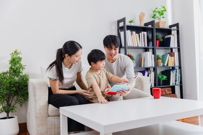 A young family sits on a couch, sharing a colorful children's book. The parents engage their child with expressions of joy in a well-lit, inviting room.