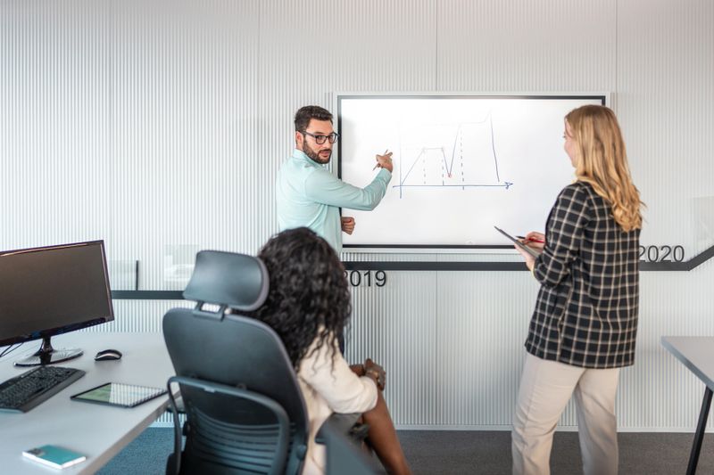 A focused mid adult Hispanic man in smart casual attire presents financial graphs on a whiteboard, engaging with his diverse, professional team in a bright office setting.