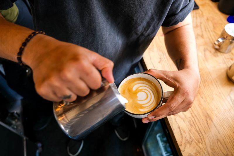 Front view male barista pouring froth milk on coffee cup prepared coffee latte art at bar counter. Step 7. Making latte art