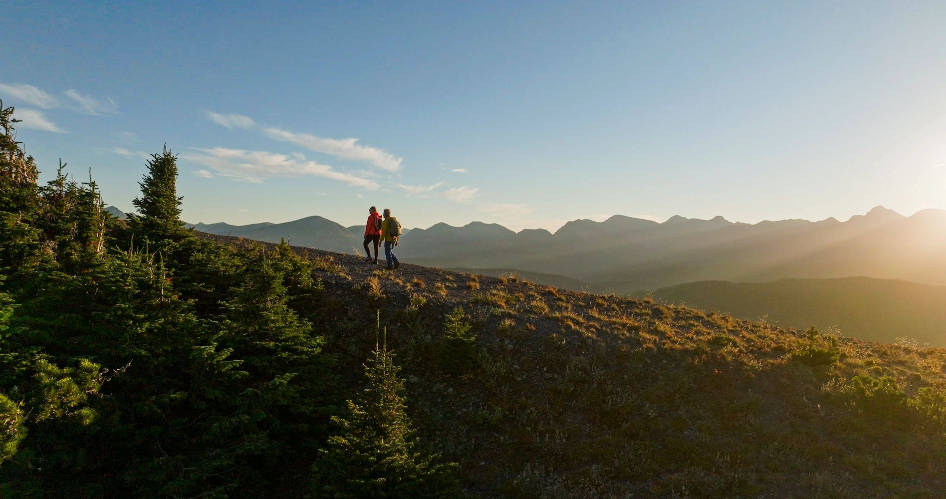 Two hikers walking on a ridge at sunrise with mountains in the background.