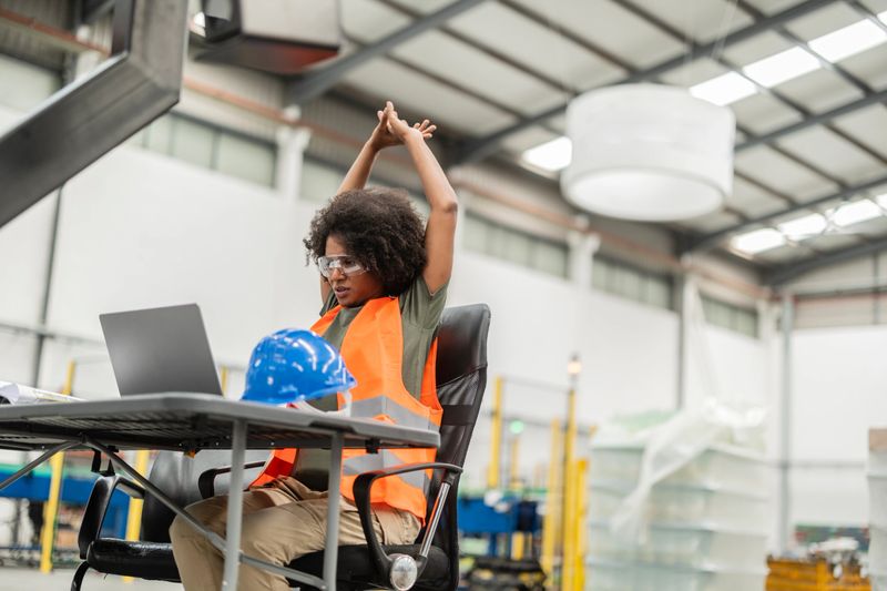 Multiracial female engineer in a robotics factory takes a break, stretching while working on a laptop in a bright, modern workspace filled with industrial equipment