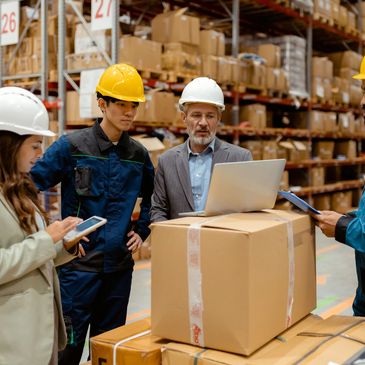 Warehouse team in hard hats reviewing inventory using laptop and tablet.