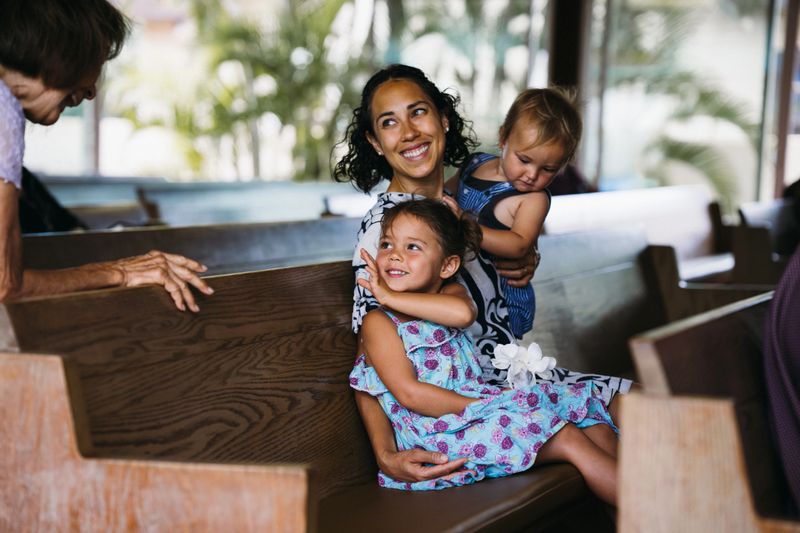 A beautiful Eurasian woman of Hawaiian and Chinese descent affectionately holds her one year old son and three year old daughter while chatting with a friend in church sanctuary before Christian religious service begins.