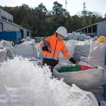 Worker in safety gear inspecting bags of plastic parts outdoors.