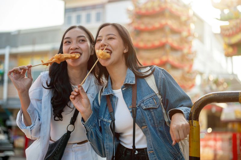 A portrait of two beautiful, joyful young Asian female tourists are enjoy street food in Thailand while exploring the city on a sunny day. Southeast Asia, vacation, trip, summer holiday