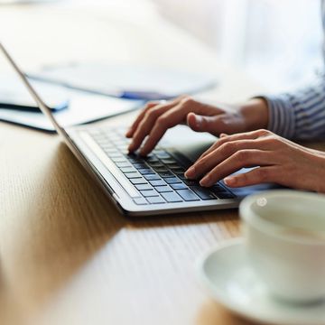 Person typing on a laptop at a wooden desk with a cup nearby.