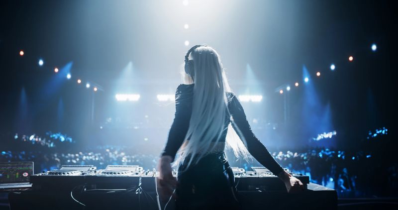 Female DJ Delivers a Live Performance of Electronic Music while Standing Behind Mixing Decks at a Massive Event. Colorful Lights and Dance Beats Play at an EDM Concert. Back View
