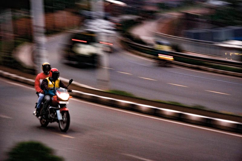 A dynamic shot capturing two motorcycle riders in motion, speeding through a bustling urban street at dusk. The image features a blurred motion effect, emphasizing the fast-paced nature of city transport. The riders are wearing protective helmets and jackets, highlighting the importance of safety while commuting. This photo encapsulates the vibrancy and energy of city life, with the road illuminated by the fading light and vehicle headlights.
