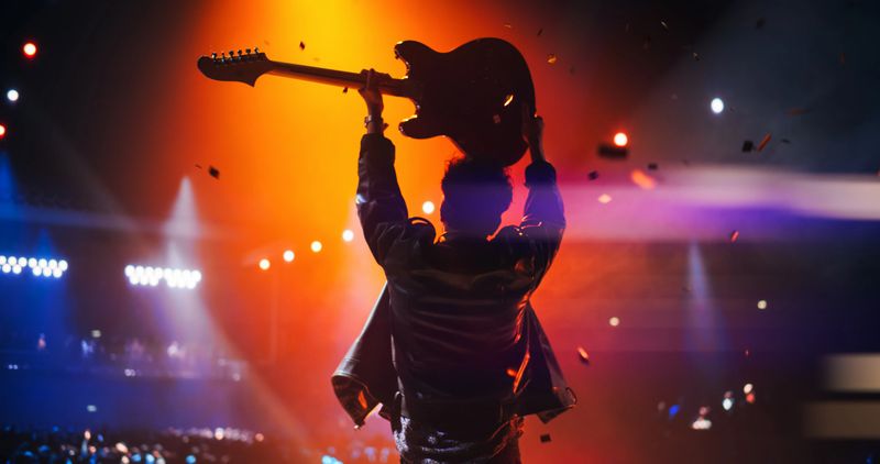 Rock Band Performing on Stage, Guitarist in a Leather Jacket and a Bandana Raising His Electric Guitar in the Air, Celebrating a Successful Concert with Devoted Audience. Footage From the Back
