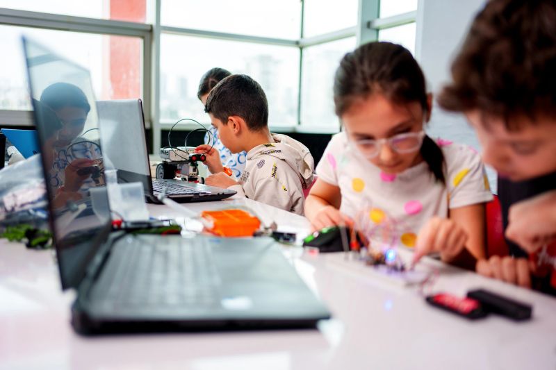 Group of young students participating in a hands-on robotics and STEM education activity. Two children are assembling a robot with various components, while others work on coding and programming using laptops. The classroom is vibrant and filled with educational tools and materials, promoting creativity, teamwork, and innovation. Perfect for themes related to technology, education, learning, and future skills development.