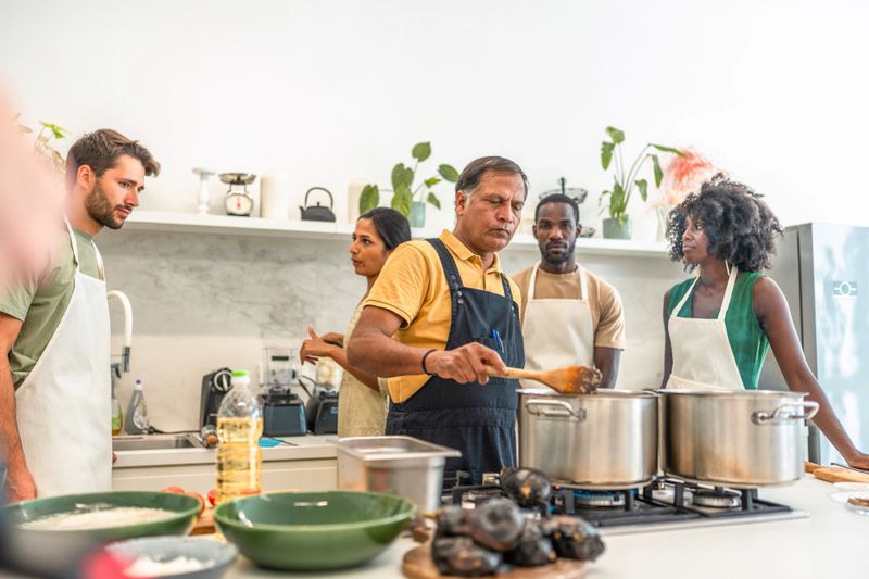 A group of diverse individuals engage in learning culinary skills with a chef guiding them in a bright, contemporary kitchen, surrounded by cooking utensils and fresh ingredients.