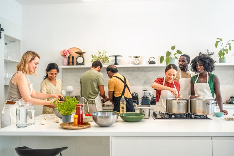 A group of cheerful attendees joyfully engage in preparing Indian cuisine, guided by an experienced chef in a modern kitchen setting, wearing casual attire and using culinary tools.