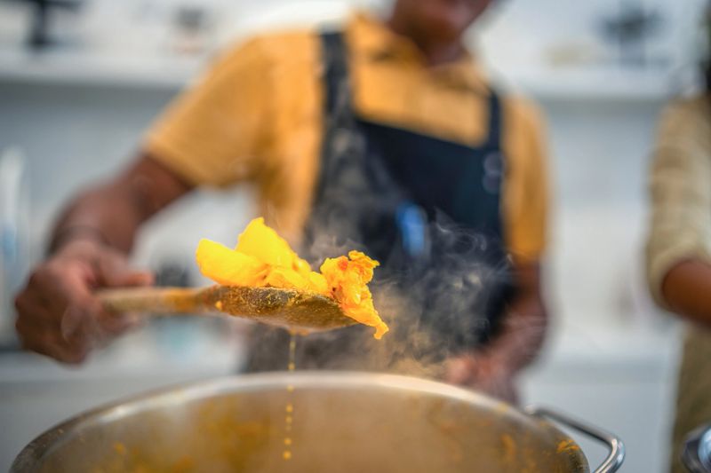 A mature chef, clad in a black apron, actively seasons a dish with vibrant spices in a well-equipped, contemporary kitchen, showcasing culinary expertise.