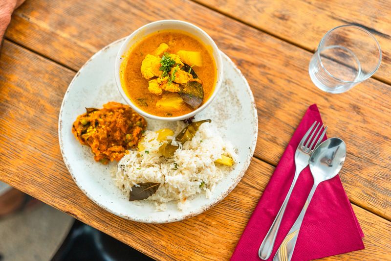 A plate of aromatic Indian food with rice, curry, and vegetable dish, prepared by an aspiring chef during a cooking class, rests on a wooden table with utensils and a glass of water.