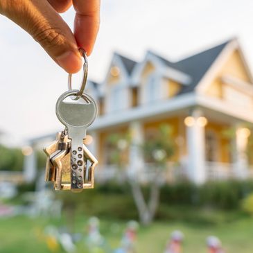 Hand holding house-shaped keys in front of a house.