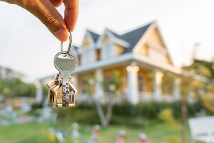 Hand holding house-shaped keys in front of a house.