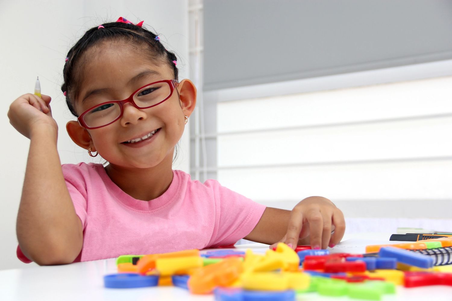 A smiling girl in red glasses plays with colorful toys at a white table.