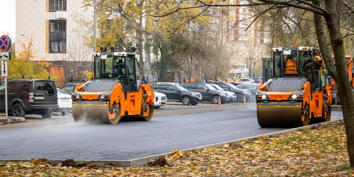 Two orange steamrollers paving a road in an urban area with autumn leaves.