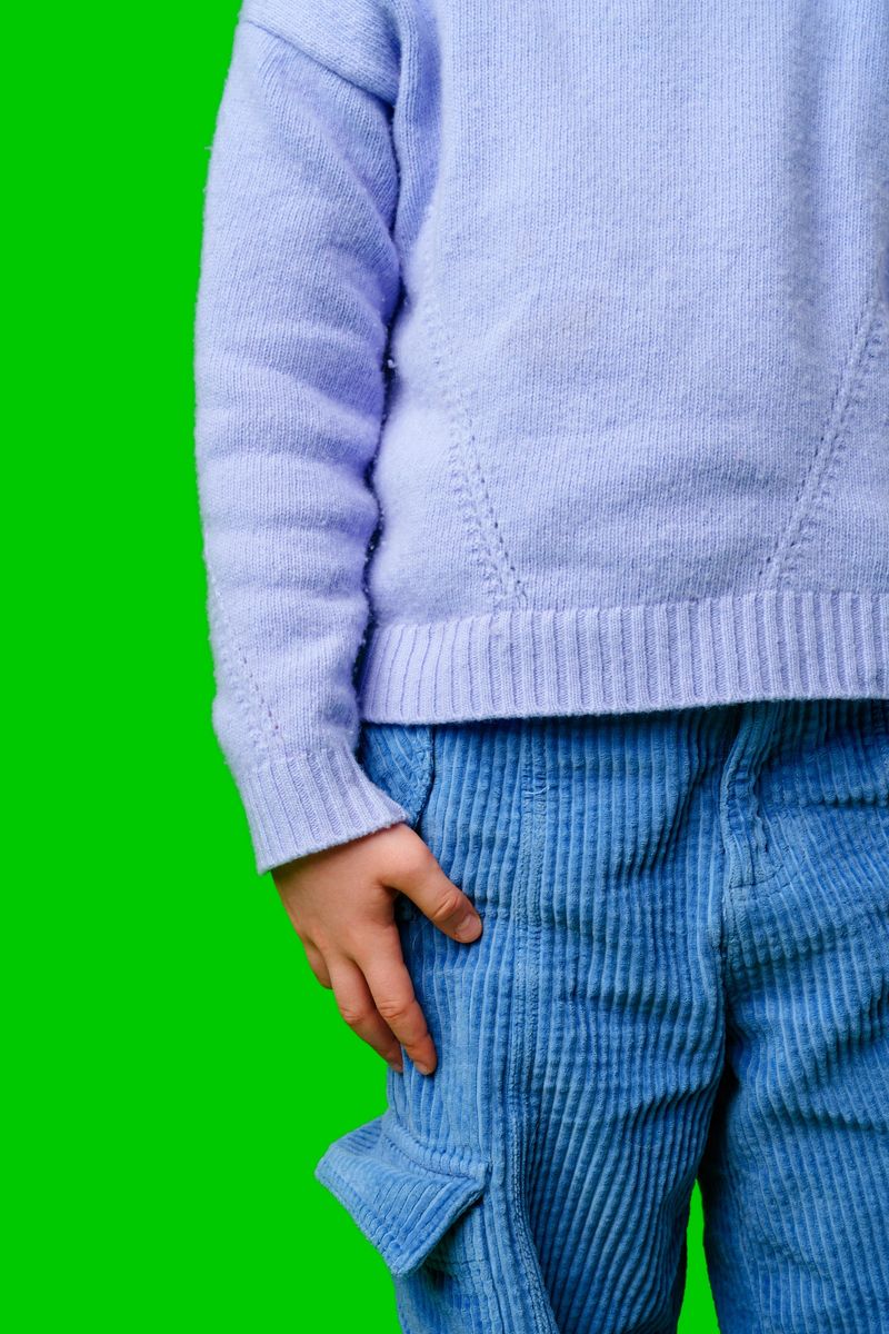 A child in a light purple sweater with blue corduroy pants stands against a vibrant green background