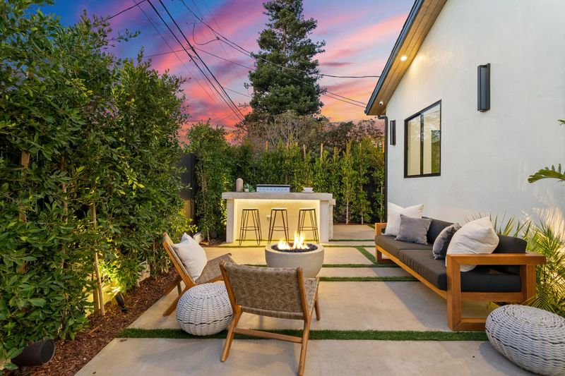 A modern outdoor patio with cozy seating around a fire pit, surrounded by lush greenery, under a vibrant sunset sky.