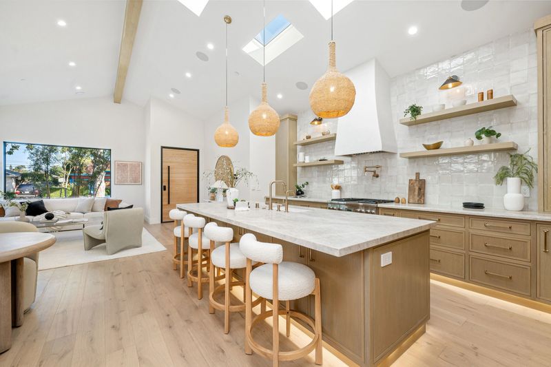 Modern kitchen interior with wooden cabinets, island, and pendant lights, featuring open shelves and a cozy living area.