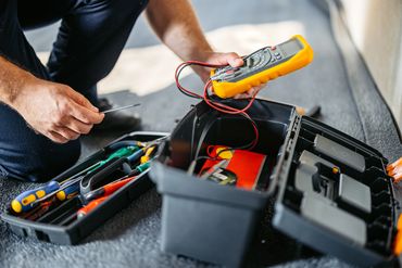 Person using a multimeter from an open toolbox filled with tools.