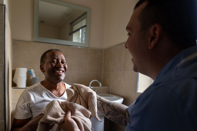 An over-the-shoulder shot of a professional male carer helping an elderly gentleman in the bathroom. The carer is drying the patient's face with a towel after a fresh shave. The two are smiling and engaging in a light-hearted moment.

Videos are available similar to this scenario.