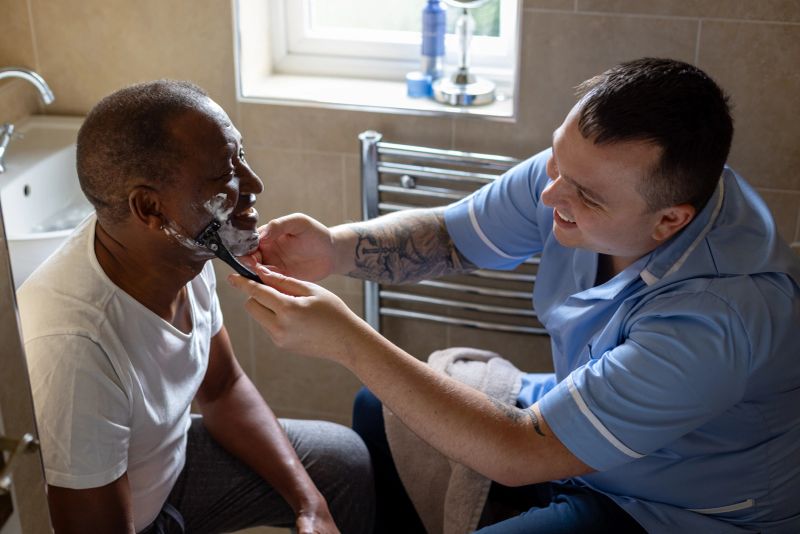 A waist-up shot of a professional male carer wearing a full uniform helping an elderly gentleman in the bathroom. The carer is shaving the patient's face with a razor. They are smiling at each other and engaging in conversation. Videos are available similar to this scenario.