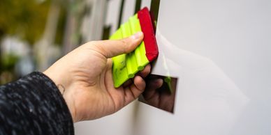 Hand using a green and red squeegee to apply vinyl lettering on a white surface.