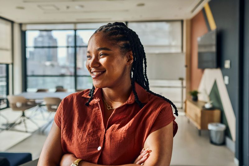 Portrait of a young black female software engineer smiling with crossed arms, enjoying her work in a modern office environment