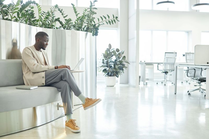 Side view of African American male entrepreneur typing on laptop working in lounge zone of coworking space featuring combination of metal accents and green plants in interior, copy space
