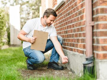 Man inspecting foundation of brick house with clipboard in hand.