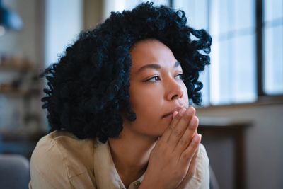 A woman with curly hair looks thoughtfully out a window, hands near her face.