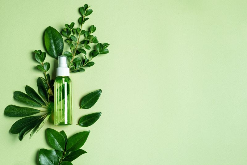 Bottle of cosmetic product over green leaves on green background, top view, copy space. Body mist, vert spray. Sustainable lifestyle, beauty spa treatment concept, organic herbal oil brand packaging mock up