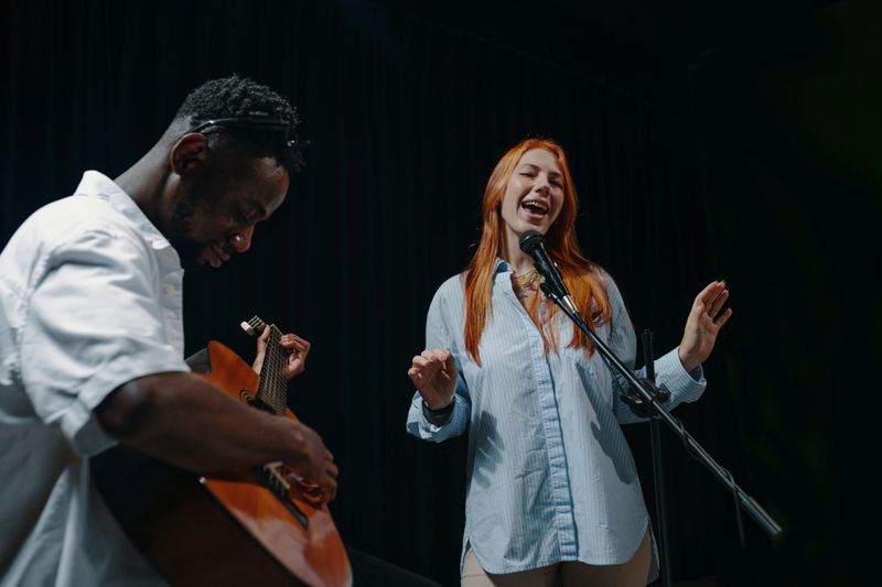 Woman singing passionately with microphone while man playing guitar on stage during performance. Focused on delivering lively show with expressions of joy and engagement