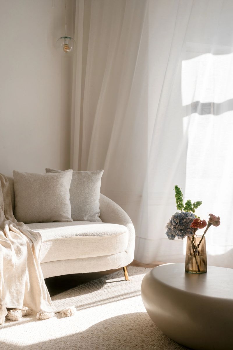 Living room features a curved couch adorned with decorative pillows, a stylish coffee table with flowers in vase and soft curtains, creating a warm atmosphere perfect for relaxation and comfort.