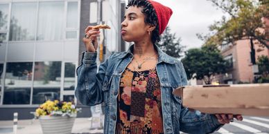 Person eating pizza slice outdoors wearing a red beanie and denim jacket.