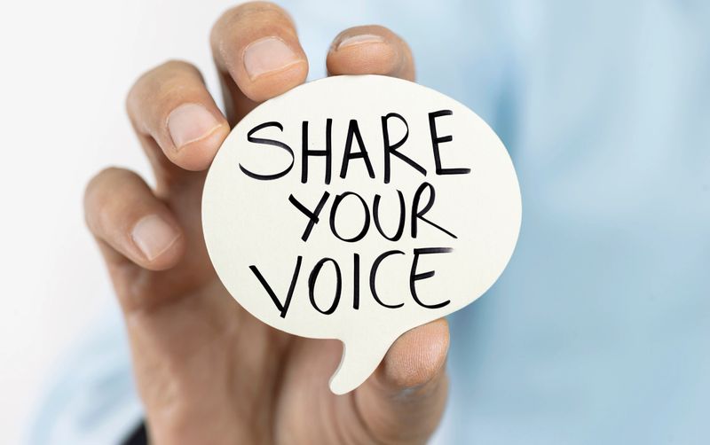 A person holding a small speech bubble cutout with the motivational message "Share Your Voice" handwritten in bold letters, set against a soft blue background.