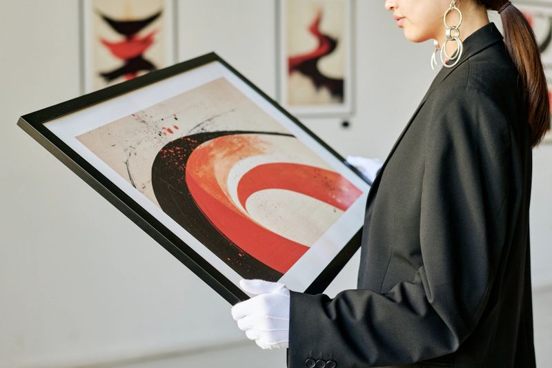 Person holding framed abstract painting in modern art gallery while examining details carefully. Exhibits and white wall in background with soft focus creating a serene atmosphere