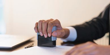 Hand stamping a document on a desk with a laptop and pen nearby.