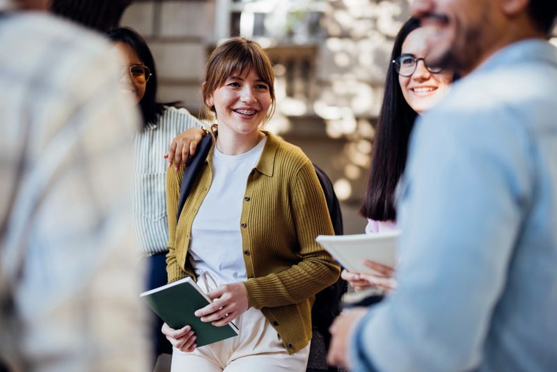A group of cheerful young students gather outside, engaging in conversation and laughter on a sunny day. They exude joy and camaraderie, creating a positive atmosphere.