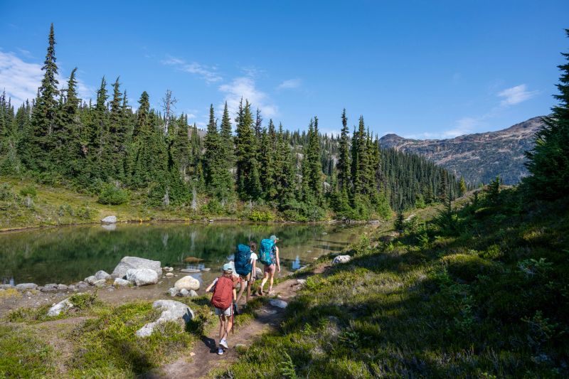 Mother and daughters backpack on mountain trail by lake, Whistler, B.C.