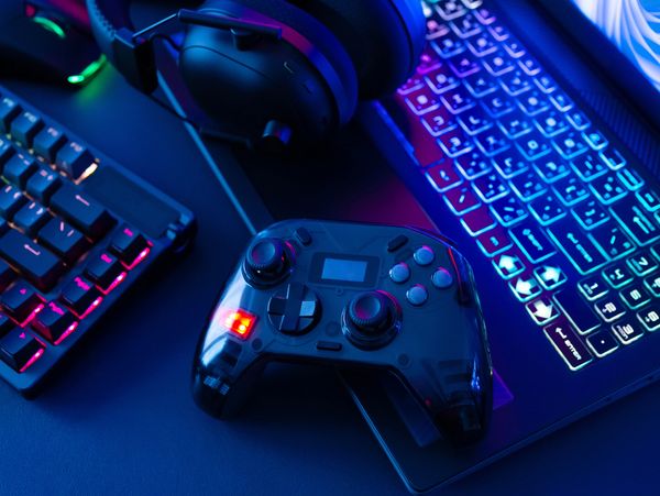 Gaming setup with controller, keyboard, headset, and laptop lit by colorful RGB lights.