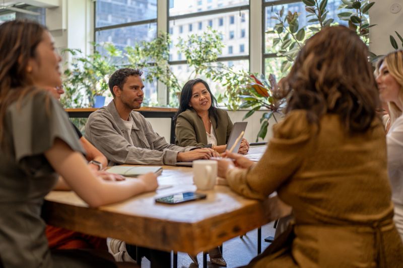 A multicultural team engaged in a collaborative discussion in a bright and modern corporate setting. The group, seated around a large wooden table surrounded by natural light and greenery, reflects inclusivity, teamwork, and innovation in a professional environment.