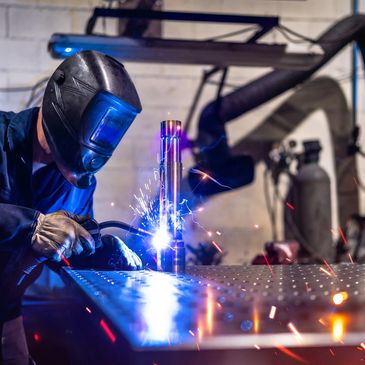 Welder in protective gear sparks bright blue welding on metal table.